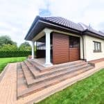 Przesuwna zabudowa tarasu lamele aluminium. Nowoczesny dom z tarasem i schodami z kamienia, otoczony zielonym ogrodem.