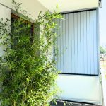 Zabudowa balkonu z lamelami pion aluminium. Zielona roślina przy nowoczesnej szarej bramie na zewnątrz.