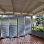 Zabudowa tarasu aluminium RAL 9006 DS. Nowoczesne aluminiowe rolety na tarasie z drewnianym podłogą i roślinami.