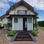 Zabudowa tarasu aluminium RAL 9006 DS. Nowoczesny dom z balkonem i tarasem, otoczony zielenią i brukowanym podjazdem.