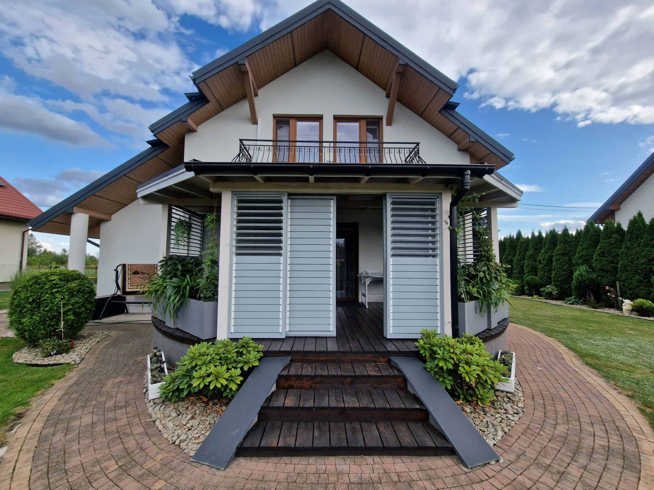 Zabudowa tarasu aluminium RAL 9006 DS. Nowoczesny dom z balkonem i tarasem, otoczony zielenią i brukowanym podjazdem.