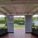 Zabudowa tarasu aluminium RAL 9006 DS. Otwarta drewniana weranda z widokiem na ogród i zielone krzewy.