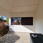 Zabudowa tarasu lamele drewnopodobne M3P20 poziome. Widok na balkon z białymi ścianami i czarnymi oknami, z widokiem na ogród.