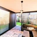 Zabudowa tarasu z lamelami aluminium pion. Przytulny balkon z meblami ogrodowymi i roślinami w tle.