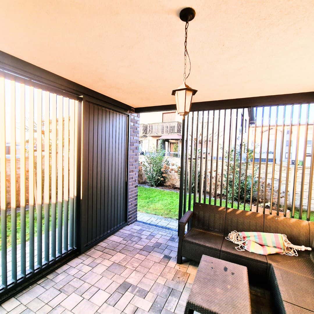 Zabudowa tarasu z lamelami aluminium pion. Przytulny balkon z meblami ogrodowymi i roślinami w tle.