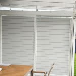 Przesuwna zabudowa tarasu z aluminium RAL 7047. Białe rolety zewnętrzne na oknach w nowoczesnym domu.