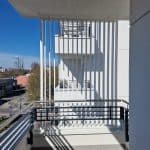 Zabudowa balkonu aluminiowa pion RAL 9016. Widok z balkonu na nowoczesny budynek i ulicę w słoneczny dzień.