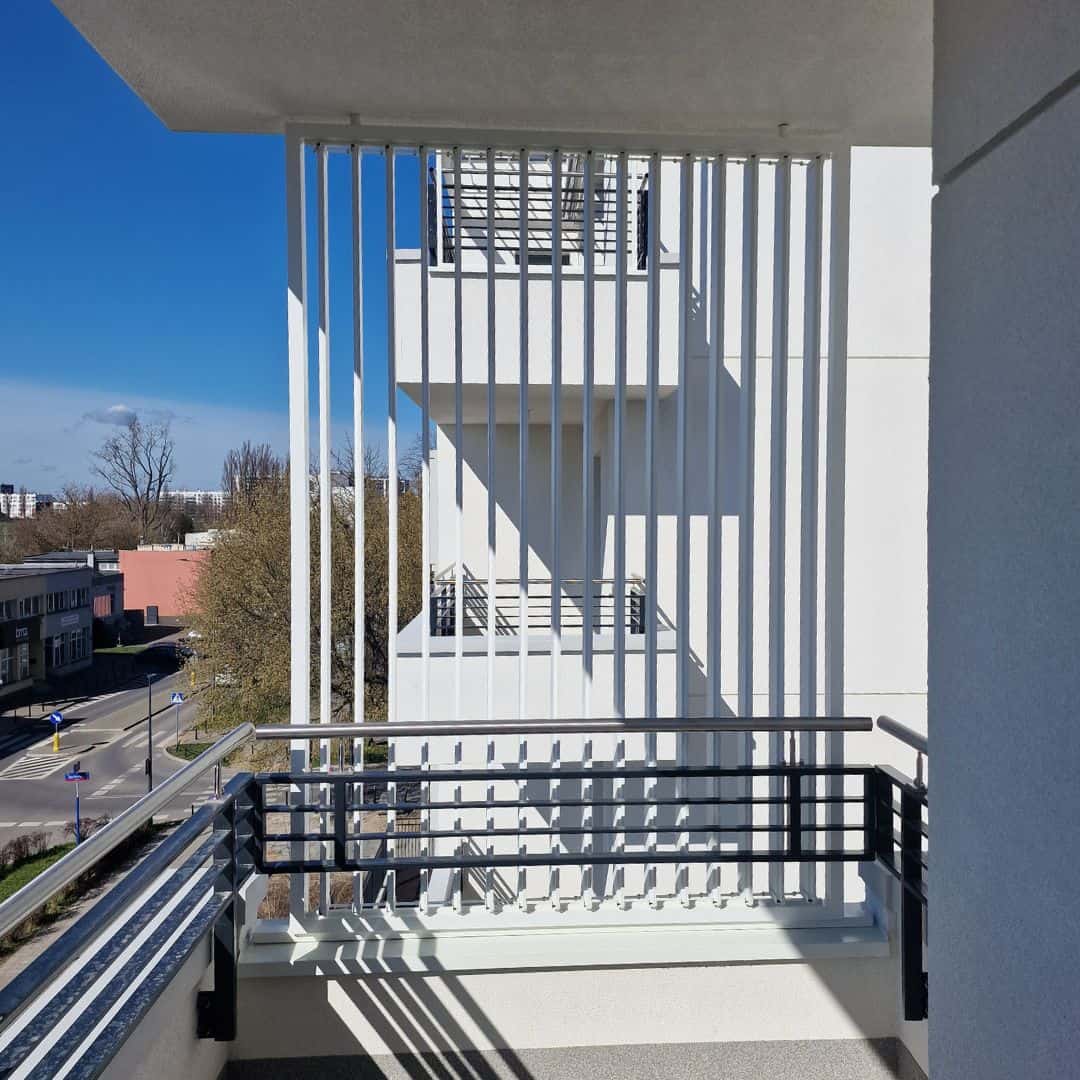 Zabudowa balkonu aluminiowa pion RAL 9016. Widok z balkonu na nowoczesny budynek i ulicę w słoneczny dzień.