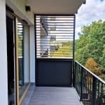 Zabudowa balkonu lamele drewnopodobne dąb D4P27. Nowoczesny balkon z drewnianymi lamelami i widokiem na zieleń.