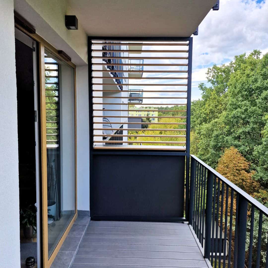 Zabudowa balkonu lamele drewnopodobne dąb D4P27. Nowoczesny balkon z drewnianymi lamelami i widokiem na zieleń.