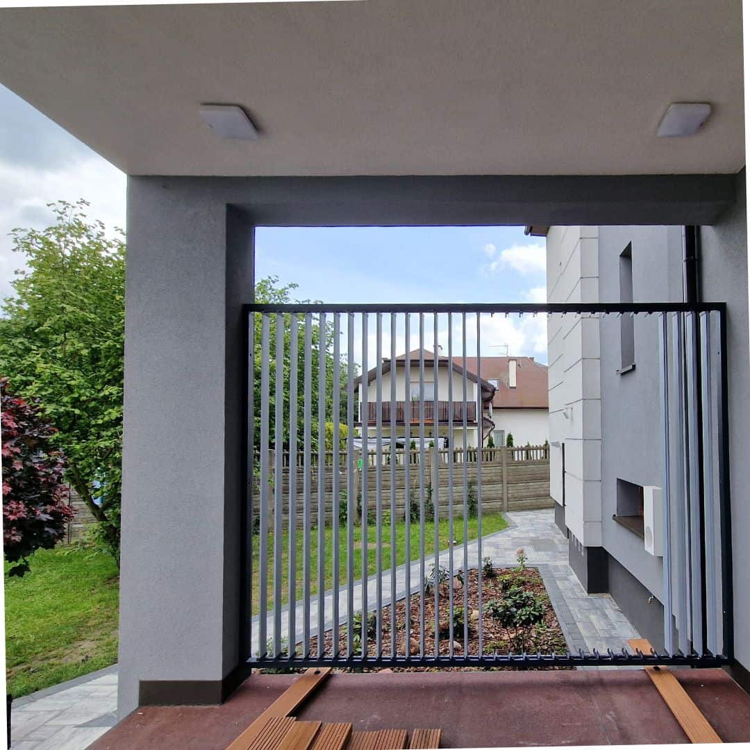Nowoczesny balkon z metalową bramą i widokiem na ogród i domy w tle.