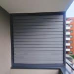 Boczna zabudowa balkonu aluminium RAL 7037. Nowoczesny żaluzja balkonowa na tle budynku mieszkalnego.